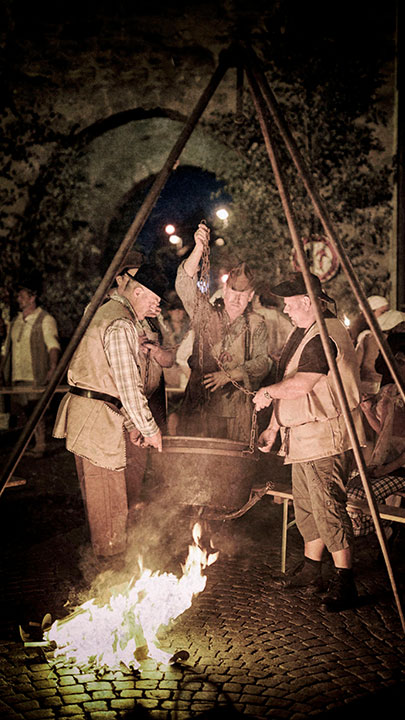 Drei Männer in historischen Kostümen kochen in einem Kessel über offenem Feuer bei einer mittelalterlichen Veranstaltung.