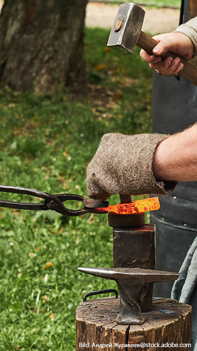 Nahaufnahme eines Schmieds, der glühendes Eisen auf einem Amboss mit einem Vorschlaghammer bearbeitet.