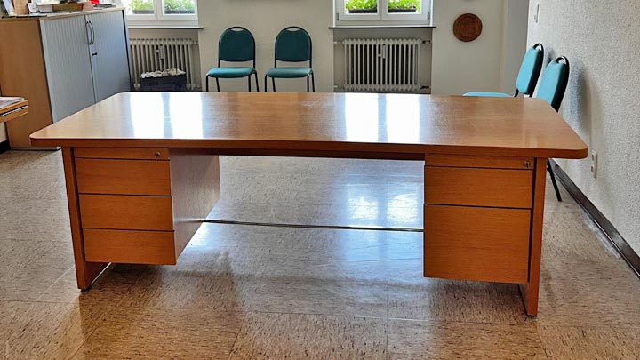 Ein großer, klassischer Holzschreibtisch mit einer glatten, hellbraunen Tischplatte, abgerundeten Ecken und zwei Schubladenschränken an den Seiten.