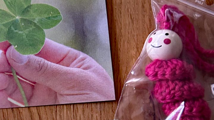 Ein Foto einer Hand, die ein vierblättriges Kleeblatt hält, neben einem gehäkelten pinken Glückswürmchen mit einem lächelnden Holzkopf in einer Plastikverpackung.