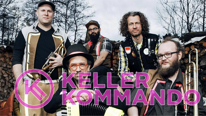 Die fünf Mitglieder der Band Kellerkommando posieren mit Instrumenten wie Trompete und Akkordeon vor einem Holzstapel. Der Bandname ist in großen, pinken Buchstaben im Vordergrund eingeblendet.