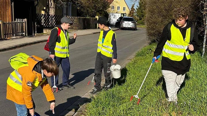 Vier Jugendliche in gelben Warnwesten sammeln mit Müllgreifern und einem Eimer Abfall im Gras an einem Straßenrand.