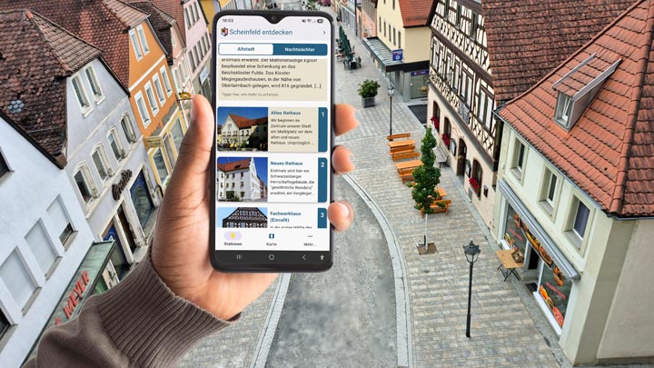 Blick auf ein Mobiltelefon mit der geöffneten App. Im Hintergrund eine Häuserzeile in der Hauptstraße Scheinfelds